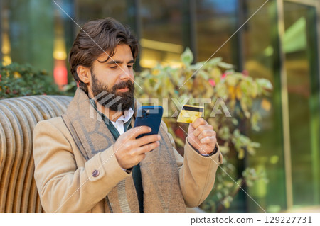 Indian man using credit bank plastic card and smartphone shopping online on downtown city street 129227731