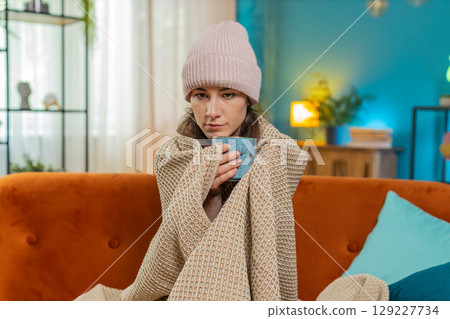 Young unhealthy sick ill woman covered by warm blanket drinks coffee freezes from cold flu at home Young unhealthy sick ill woman covered by warm blanket drinks coffee freezes from cold flu at home 129227734