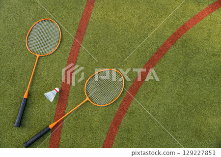 Two orange badminton rackets and a shuttlecock rest on a green sports field, captured from above. Two orange badminton rackets and a shuttlecock rest on a green sports field, captured from above. 129227851