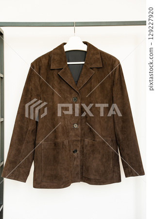Dark brown suede jacket with buttons and patch pockets hanging on white hanger in bright room 129227920