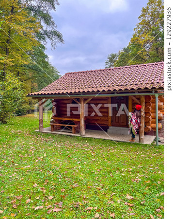 Woman in bright red cap and checkered blanket stands near wooden rustic cabin with cup in morning Woman in bright red cap and checkered blanket stands near wooden rustic cabin with cup in morning 129227936