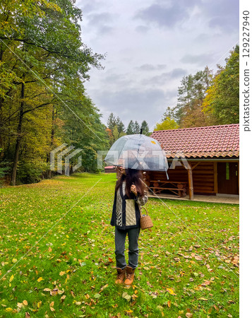 A woman stands under a transparent umbrella on a rainy autumn day in a forest near a wooden cabin A woman stands under a transparent umbrella on a rainy autumn day in a forest near a wooden cabin 129227940