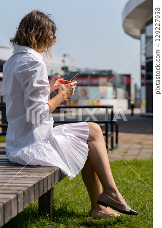 Woman using smartphone and holding red disposable cup while sitting on a bench outdoors in summer 129227958