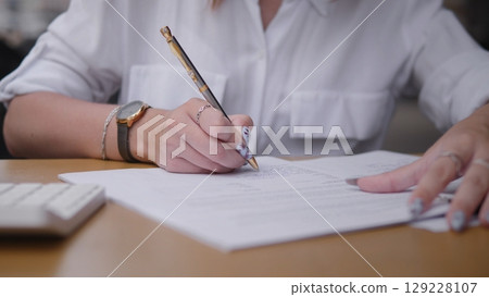 An individual diligently jotting down notes on paper with a pen at a cluttered desk full of papers and items 129228107