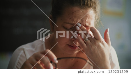 Exhausted Female Teacher Taking Off Glasses, Sitting Alone in Empty Classroom, Feeling Stress Exhausted Female Teacher Taking Off Glasses, Sitting Alone in Empty Classroom, Feeling Stress 129231299