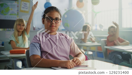 Pretty Primary School Girl Sitting at the Desk, Smiling and Looking at Camera in Colorful Classroom Pretty Primary School Girl Sitting at the Desk, Smiling and Looking at Camera in Colorful Classroom 129231400