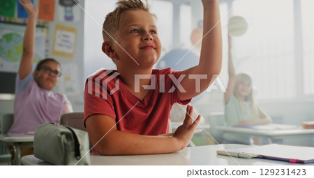 Elementary School Boy Sitting at Desk, Raising Hand and Answering a Teacher's Question 129231423