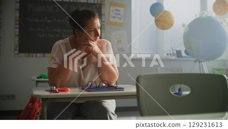 Tired Female Teacher Sitting Alone at the Desk in Empty Classroom, Relaxing After Class 129231613