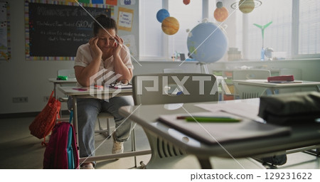 Exhausted Female Teacher Rubbing her Temples, Sitting Alone at Desk in Empty Classroom 129231622