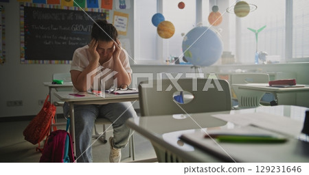 Exhausted Female Teacher Rubbing her Temples, Sitting Alone at Desk in Empty Classroom Exhausted Female Teacher Rubbing her Temples, Sitting Alone at Desk in Empty Classroom 129231646
