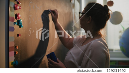 Female Teacher Writing Numbers and Multiplication Table on the Chalkboard, Teaching Primary School Students 129231692