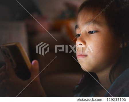 Asian girl using smartphone at night with glowing screen, concept of screen addiction and harmful blue light in children 129233100