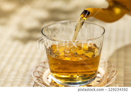 Pouring barley tea from a kettle into a glass on a tatami mat gives you a cool, Japanese summer feeling. Pouring barley tea from a kettle into a glass on a tatami mat gives you a cool, Japanese summer feeling. 129234392