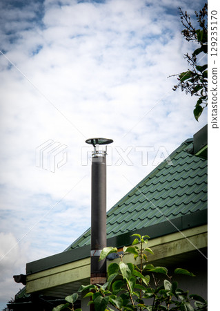 Modern Chimney Pipe on Green Metal Roof Against Cloudy Sky. 129235170