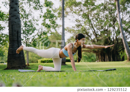 Asian woman practicing yoga outdoors, performing bird-dog pose on a yoga mat in a green park 129235512