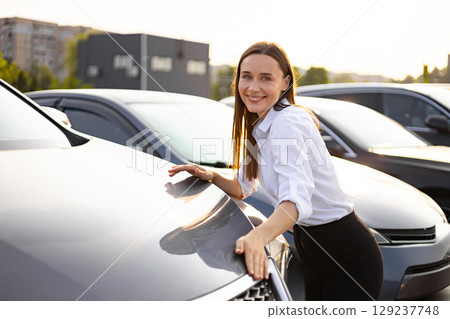 A smiling woman leans against a car, appearing excited about a potential purchase. A smiling woman leans against a car, appearing excited about a potential purchase. 129237748