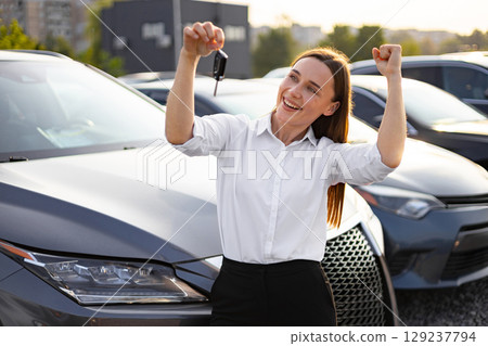 A happy woman celebrates the purchase of her new car, holding up the keys with joy at a dealership. 129237794