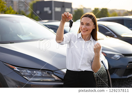 A happy woman holding car keys, celebrating her new car purchase, smiling and looking excited in front of a car dealership. 129237811