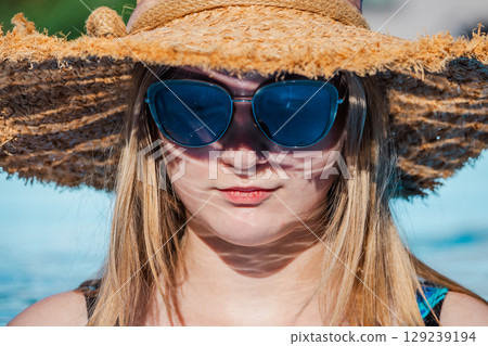 Close-up portrait of a young woman wearing a straw hat and blue sunglasses by the pool, enjoying a sunny summer day outdoors. Close-up portrait of a young woman wearing a straw hat and blue sunglasses by the pool, enjoying a sunny summer day outdoors. 129239194