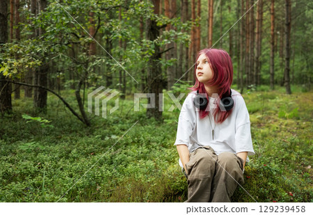 Youngster with pink hair and headphones sitting in lush forest Youngster with pink hair and headphones sitting in lush forest 129239458