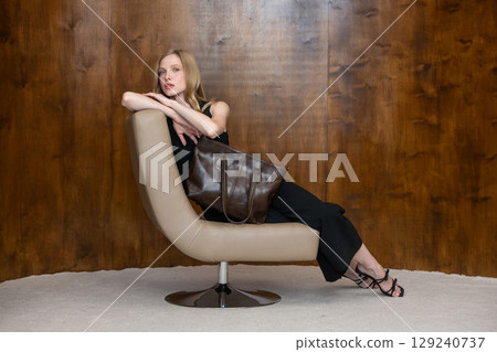 woman with wavy hair seated in a beige chair posing with a rich, dark brown leather tote bag. wearing sleeveless black top, trousers that drape gracefully to just above her black heeled sandals. 129240737