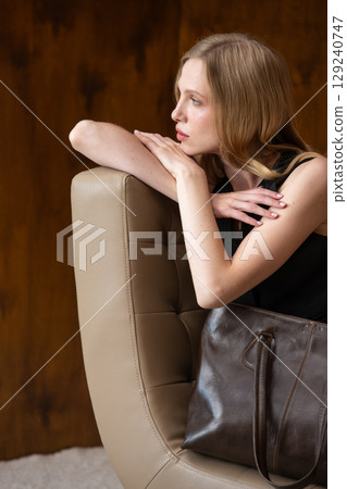 woman with wavy hair seated in a beige chair posing with a rich, dark brown leather tote bag. wearing sleeveless black top, trousers 129240747