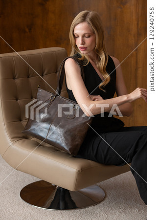 woman with wavy hair seated in a beige chair posing with a rich, dark brown leather tote bag. wearing sleeveless black top, trousers 129240758
