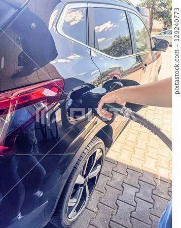 Close-up of a hand holding a fuel nozzle while filling a modern black car at a gas station. Great concept image for transport, travel, energy, and automotive themes. High quality photo 129240773