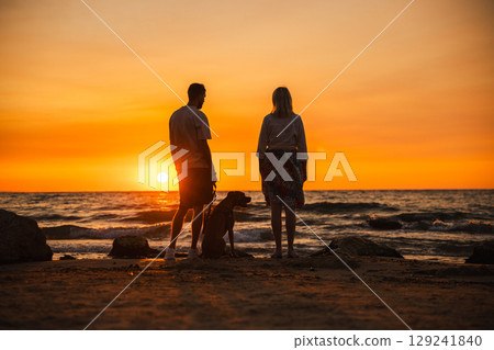 Man and woman with a German Boxer dog standing on a sandy beach at sunset, watching the sun set over the calm sea waves. 129241840