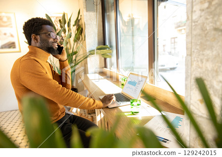 Smiling African American man wearing glasses and sweater working on laptop while talking on smartphone Smiling African American man wearing glasses and sweater working on laptop while talking on smartphone 129242015