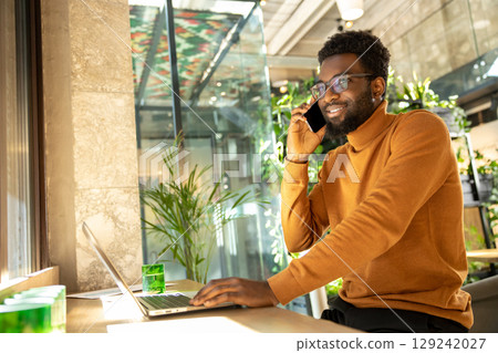 Happy man working remotely on laptop while having business phone call in bright stylish office Happy man working remotely on laptop while having business phone call in bright stylish office 129242027