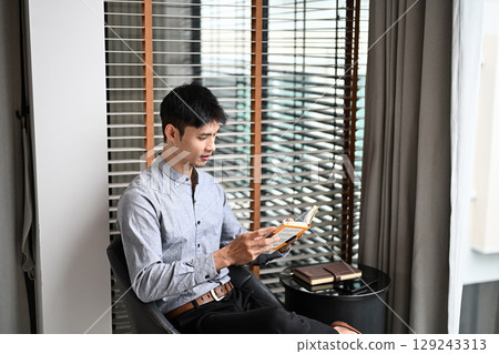 Adult businessman sitting by window reading a book in modern living space Adult businessman sitting by window reading a book in modern living space 129243313