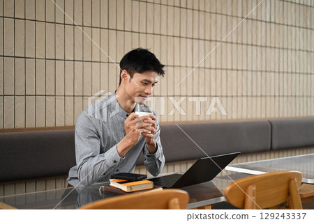 Young man enjoying coffee while working on a laptop at a modern cafe Young man enjoying coffee while working on a laptop at a modern cafe 129243337