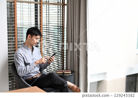 Asian businessman relaxing in office chair by the window and using smartphone Asian businessman relaxing in office chair by the window and using smartphone 129243382