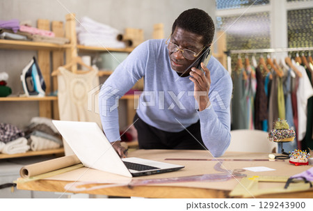 Male tailor talking on smartphone - taking order for sewing clothes in sewing workshop and entering data into laptop 129243900