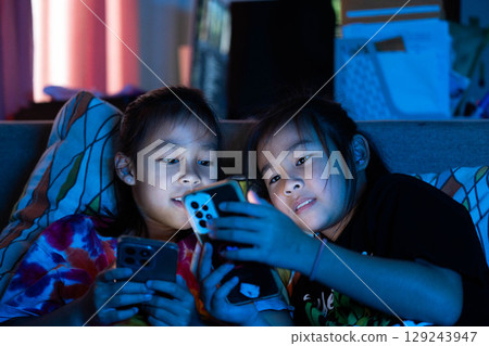 Two Asian child lying in bed using smartphone at night, screen light illuminating face, concept of screen time and digital addiction in kids Two Asian child lying in bed using smartphone at night, screen light illuminating face, concept of screen time and digital addiction in kids 129243947