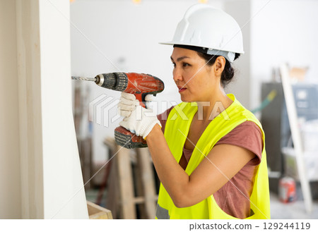 Woman builder using drill to bore hole in wall 129244119