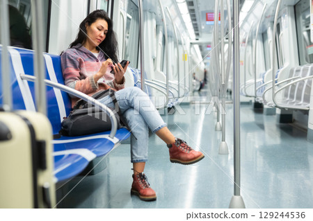 Woman with suitcase sitting in subway car and using her smartphone Woman with suitcase sitting in subway car and using her smartphone 129244536
