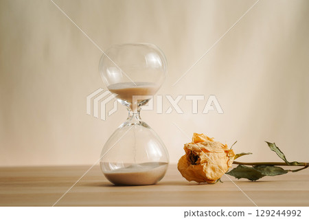 A simple hourglass on a wooden table with dry rose a beige backdrop, symbolizing time flow, aging, and minimalistic living. Represents timeless themes in a creative presentation for stylish concepts. 129244992
