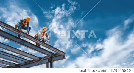Two construction workers in safety vests and hard hats work on steel roof structure under blue sky with clouds Two construction workers in safety vests and hard hats work on steel roof structure under blue sky with clouds 129245840