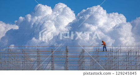 Worker in high visibility gear stands on metal truss system against backdrop of dramatic clouds 129245845