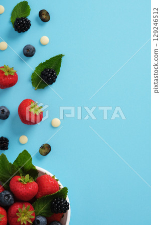 Fragment of white bowl with strawberries, blueberries, blackberries, mint leaves, and white chocolate on blue background. Space for text. 129246512