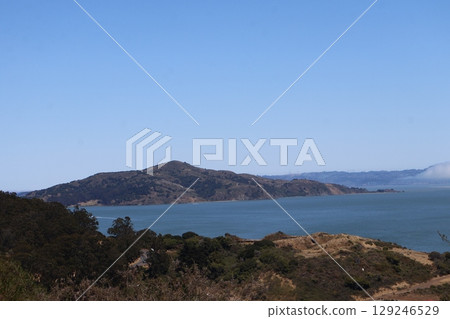 A view of the opposite shore with blue sky, blue sea bay, green forest, road, dry land, island and distant cityscape. 129246529