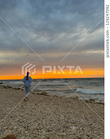 Woman walking on a sea beach against the background of the sunset Woman walking on a sea beach against the background of the sunset 129247092