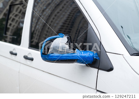 Broken side mirror of a white van temporarily fixed with blue tape, close-up on urban street background 129247211