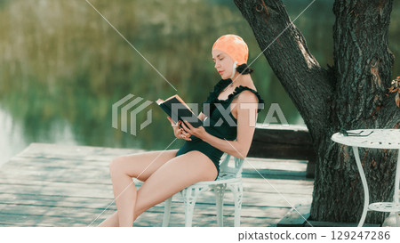 Woman in retro swimsuit reading book alone on chair near lake 129247286