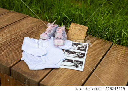 Pink baby shoes, bodysuit, ultrasound scan images, and wooden sign are laying on a wooden surface with green grass in the background, composing a sweet pregnancy announcement. 129247319
