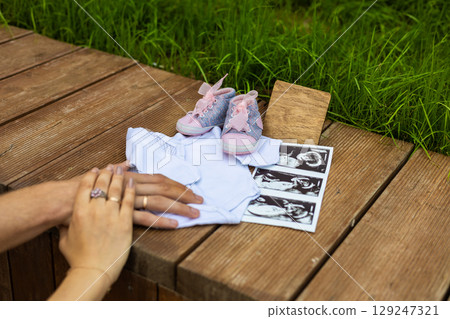 Pregnancy announcement. Baby clothes, ultrasound, tiny shoes and a wooden sign resting on wooden boards as the parents-to-be hold hands, eagerly awaiting the arrival of their little one. 129247321