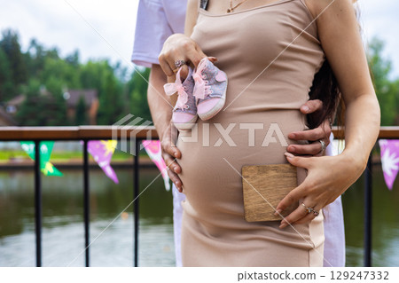 Expecting parents tenderly embrace, holding small baby shoes and a blank wooden sign against the woman's pregnant belly, symbolizing the anticipation and joy of their growing family. 129247332