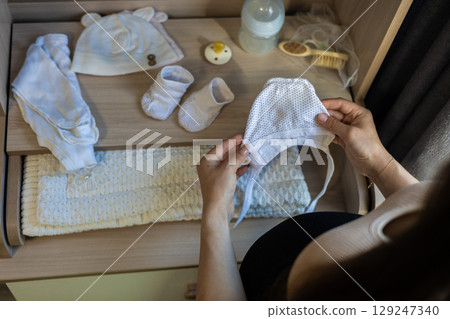Pregnant woman holding baby hat while arranging clothes and accessories in drawer, preparing for childbirth and showing love and care for her unborn child. Preparing for motherhood. 129247340